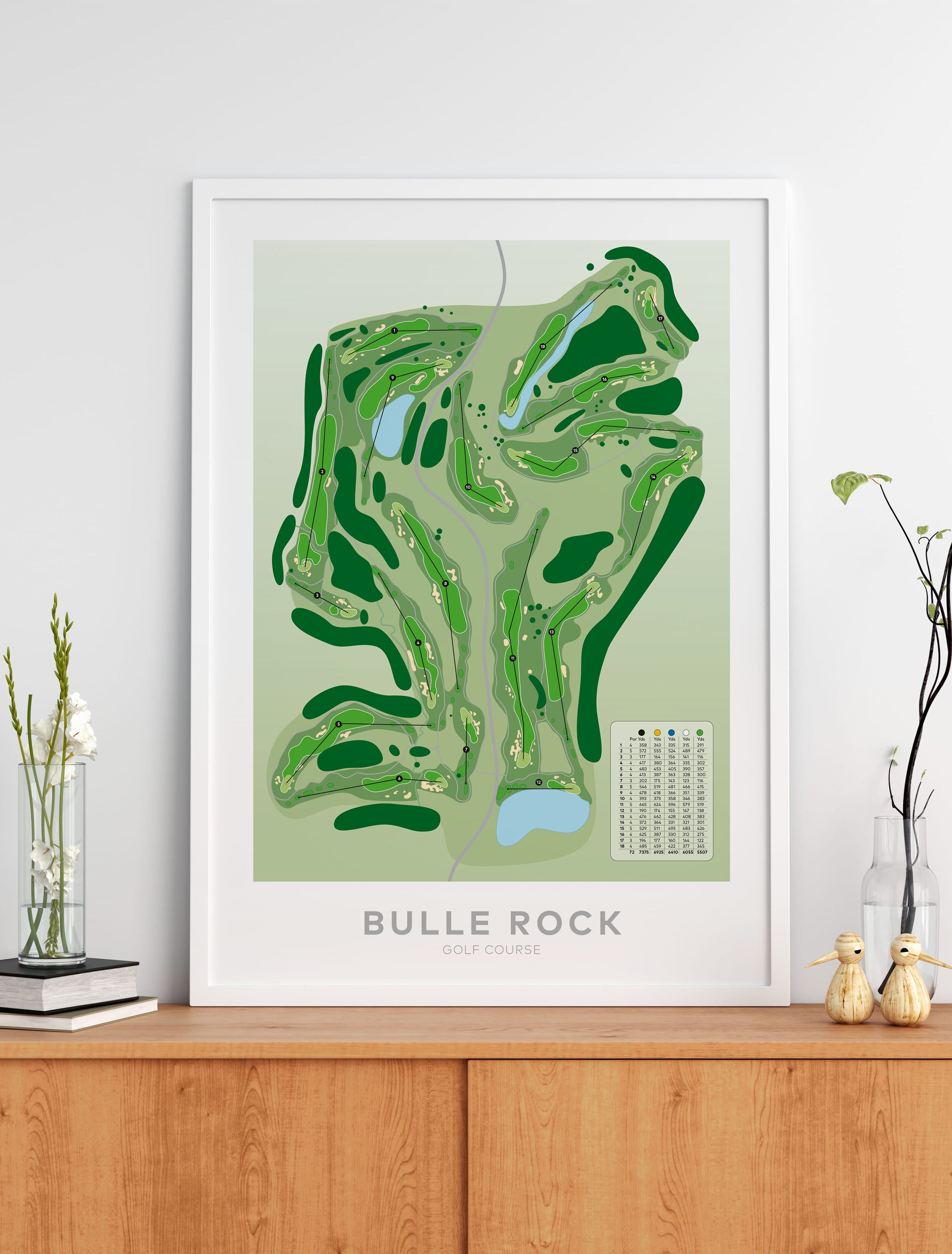 A framed golf course map print of Bulle Rock Golf Course, featuring green and blue course details on a pale green background, displayed on a wooden cabinet against a light gray wall with decorative vases.
