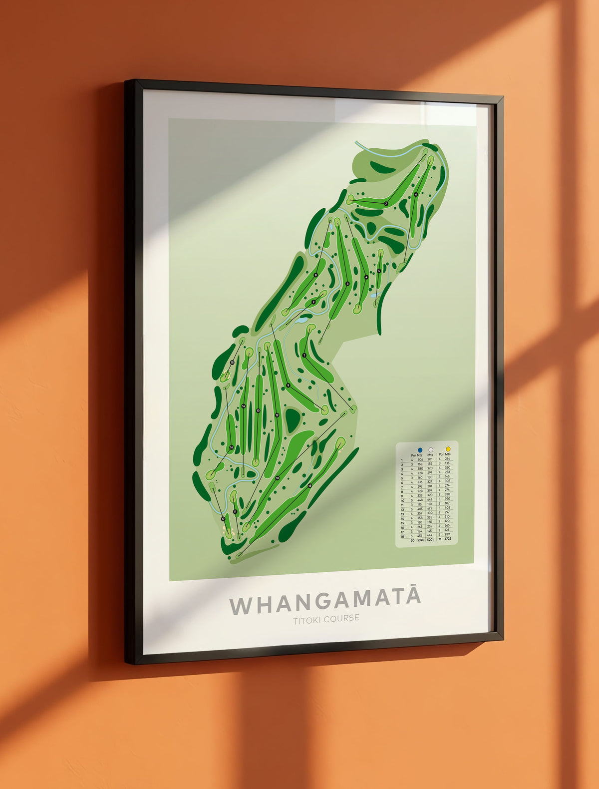 Black framed art print featuring a stylized green map of Whangamata, New Zealand, displayed on a warm terracotta wall with diagonal window light shadows.