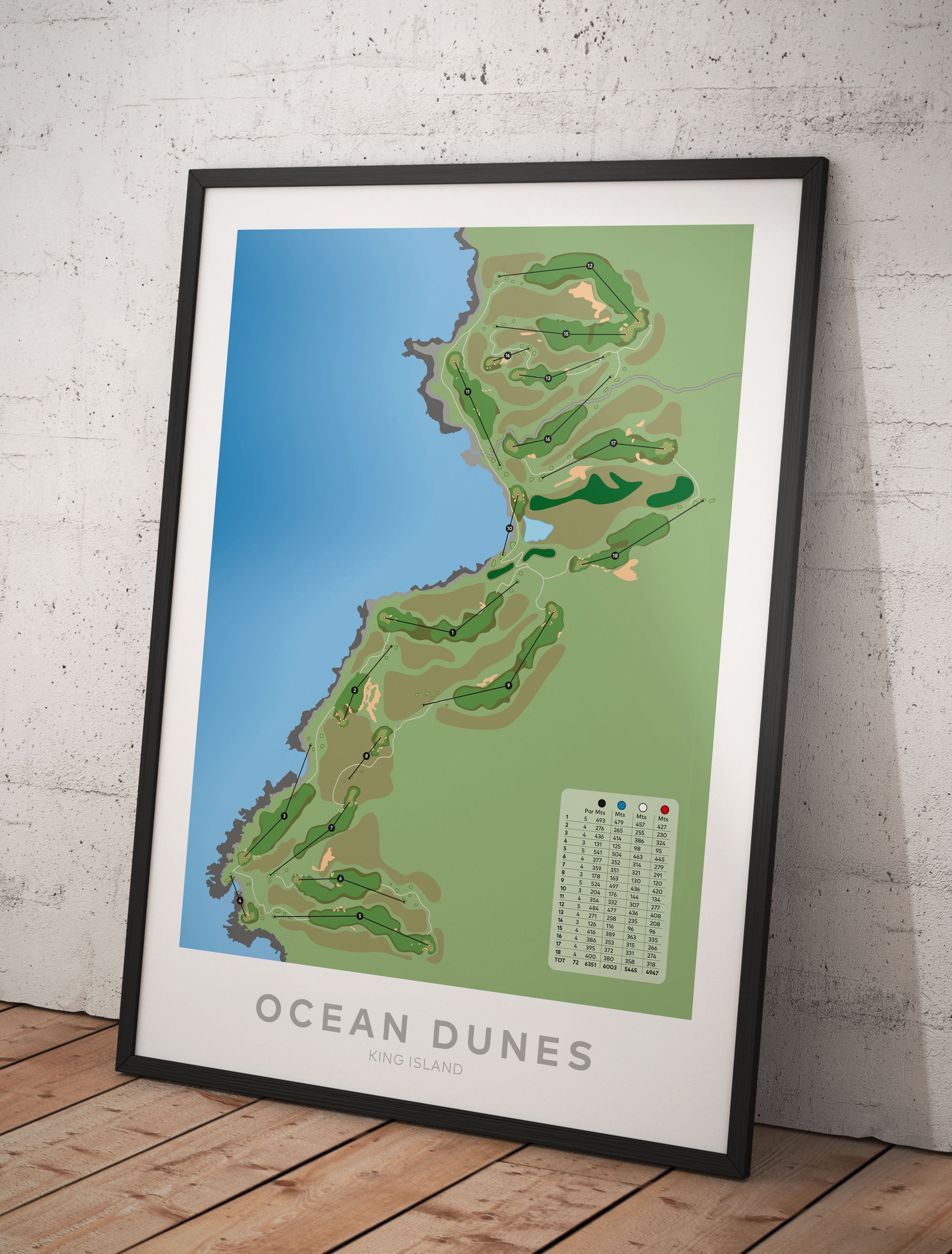 A framed golf course map poster titled 'OCEAN DUNES' leaning against a light gray brick wall on a wooden floor. The print shows a stylized map with blue water, green and tan land, and a small legend in the lower right.-australia-golf-art