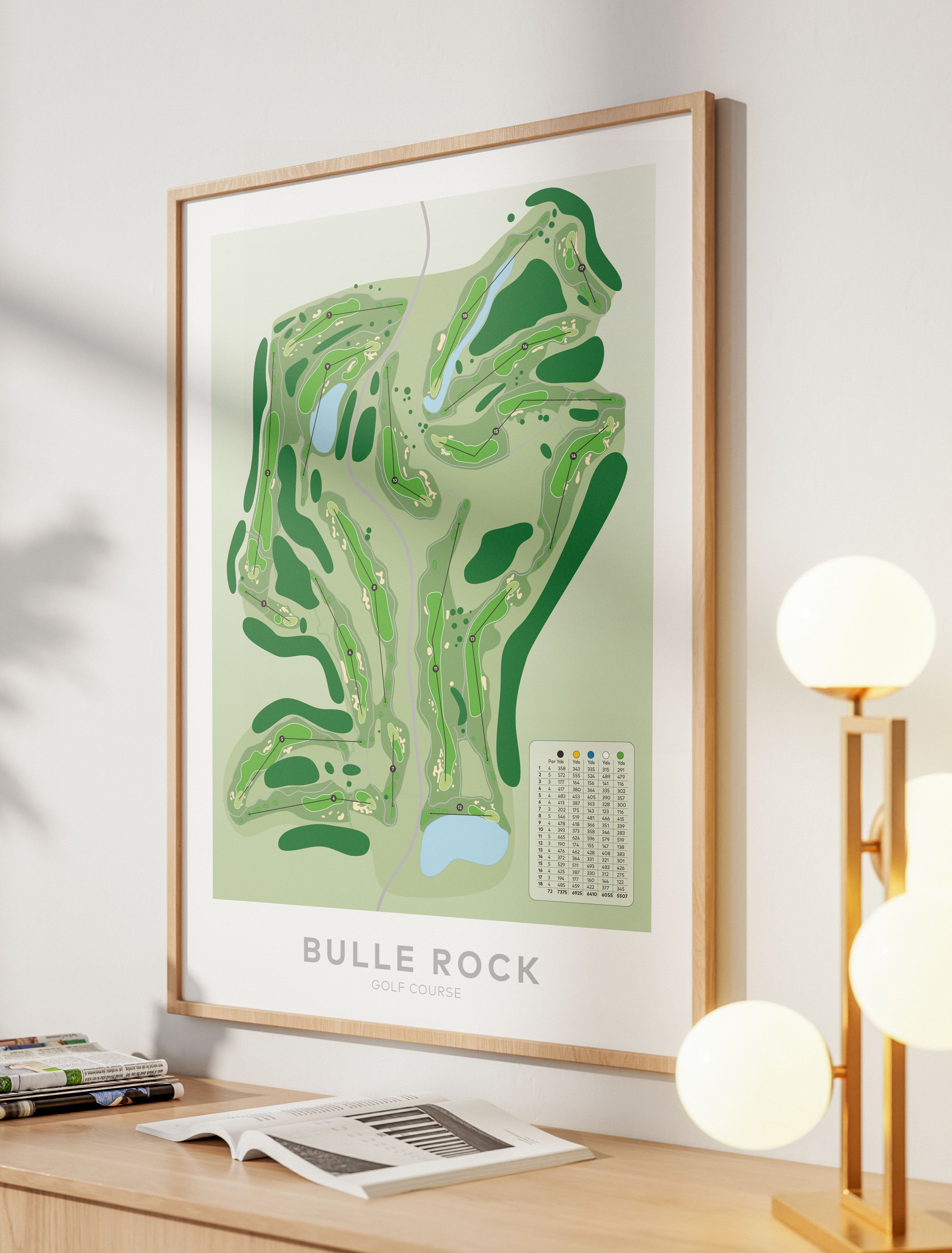 A framed golf course map print of Bulle Rock Golf Course, displayed on a wooden console table in a modern room setting with a lamp and books.