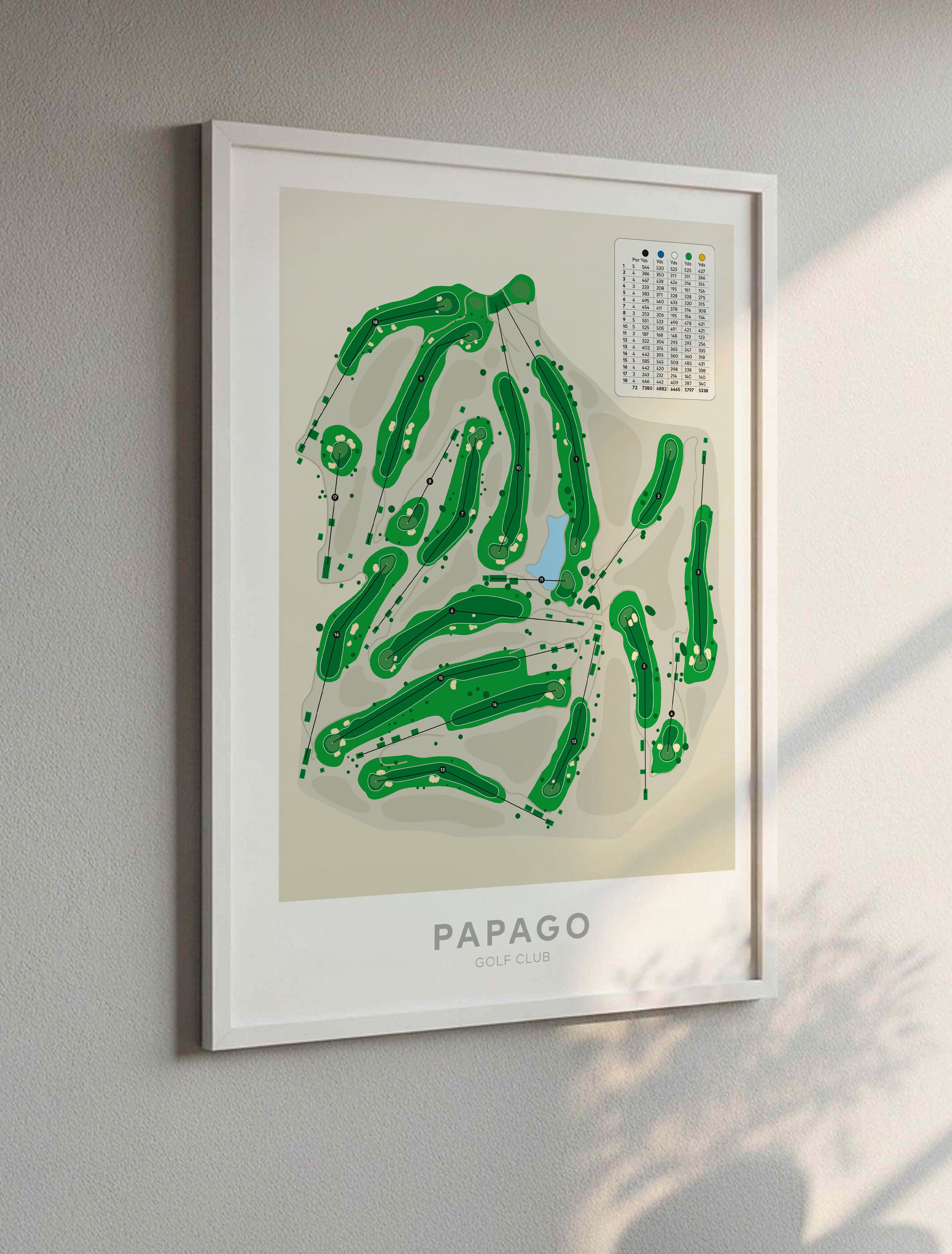 White framed art print of the Papago Golf Club layout, featuring green tee shapes on a cream background with a white frame, mounted on a gray wall with soft sunlight and shadow patterns.