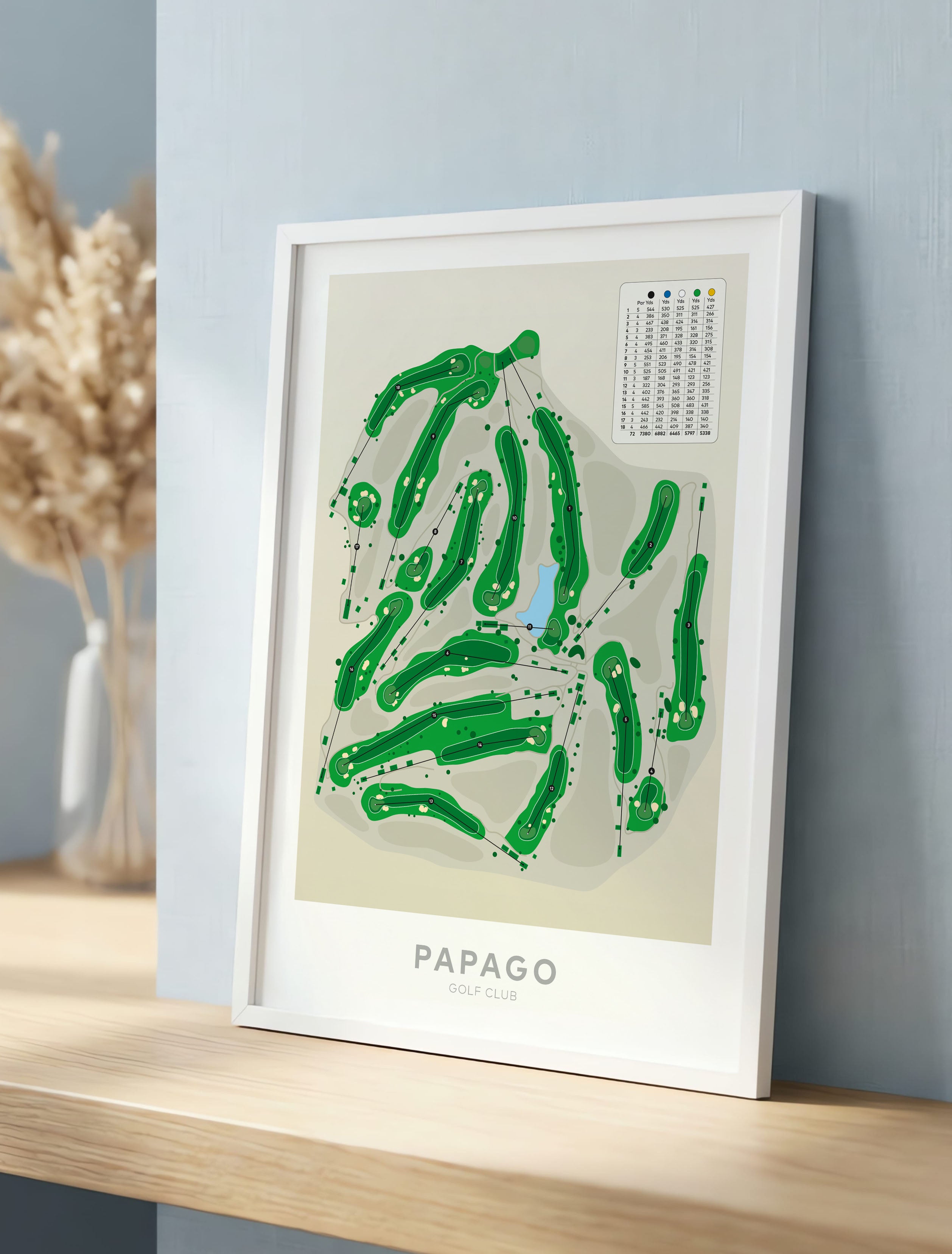 White framed golf course map print leaning against a light blue wall on a wooden shelf, with dried grass decor in the background. The print shows green course paths on a beige field with the text 'PAPAGO GOLF CLUB' at the bottom.